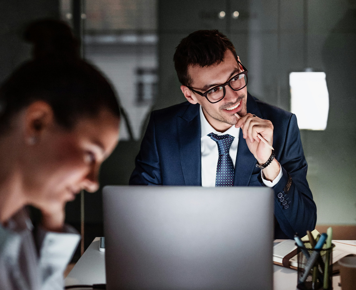 A professional man in a blue suit and glasses sits at a desk in a modern office at night. He is looking thoughtfully to the side while holding a pen to his chin. A laptop is open in the foreground, and a blurred colleague is visible in the lower-left corner.